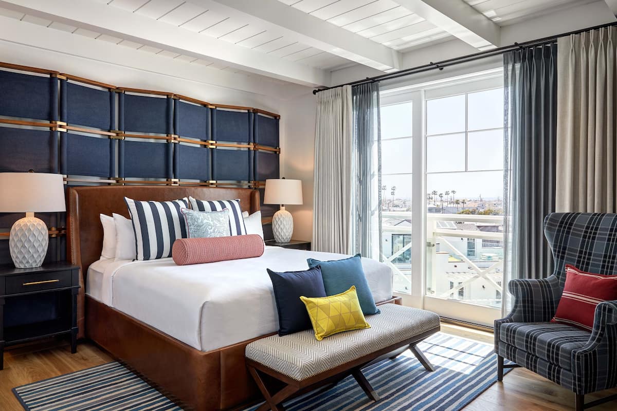 A hotel room with a large bed, a bench, a chair, and a view of the city through the window.