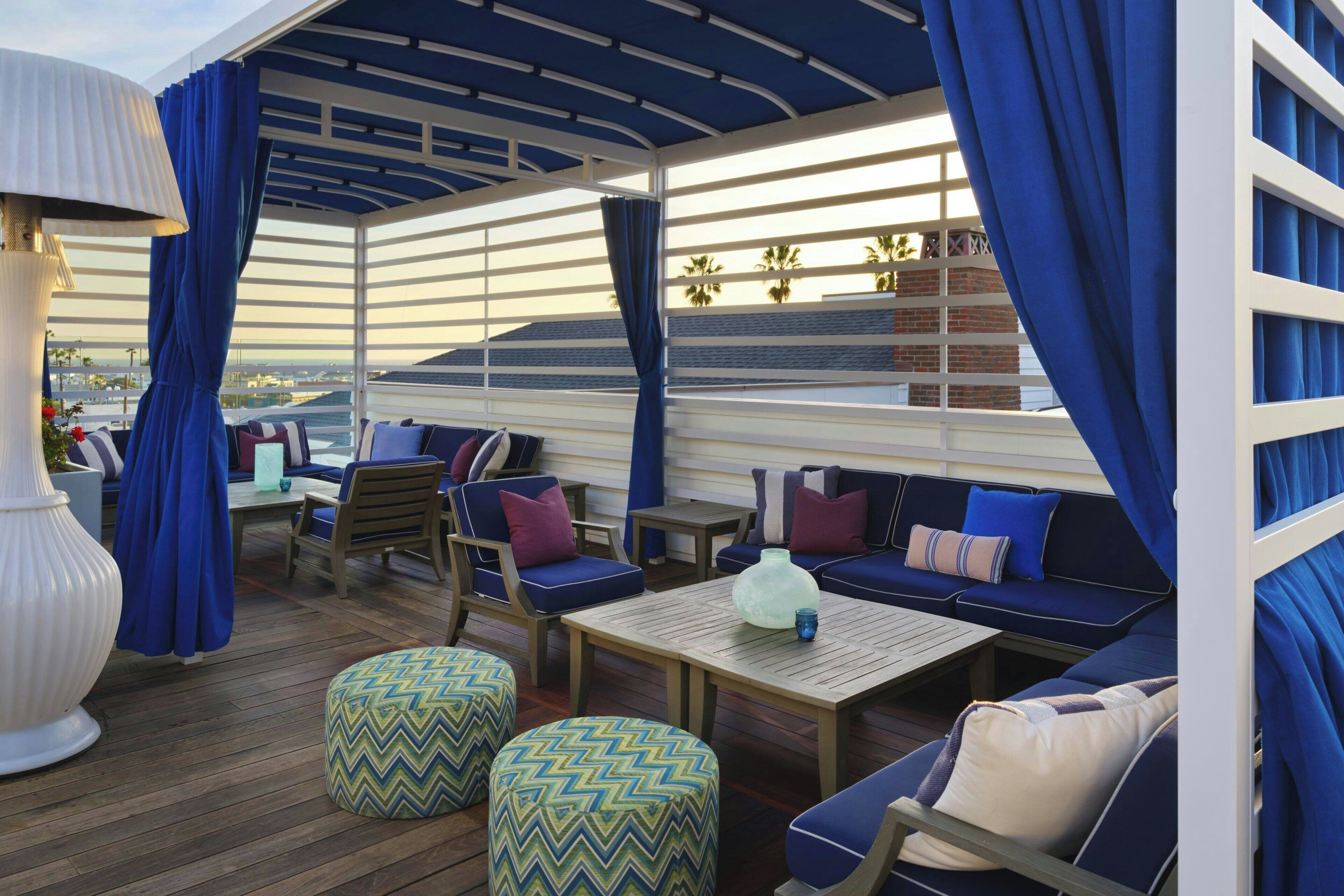 Una terraza exterior con mobiliario azul y blanco, incluyendo sofás, sillas y otomanas, bajo una zona cubierta con cortinas azules.