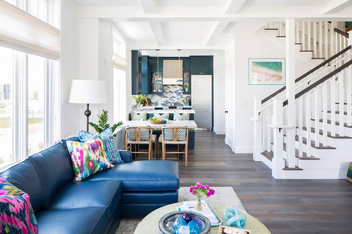 A modern living room with a blue sectional sofa, a dining area with a table and chairs, and a staircase with white railings.