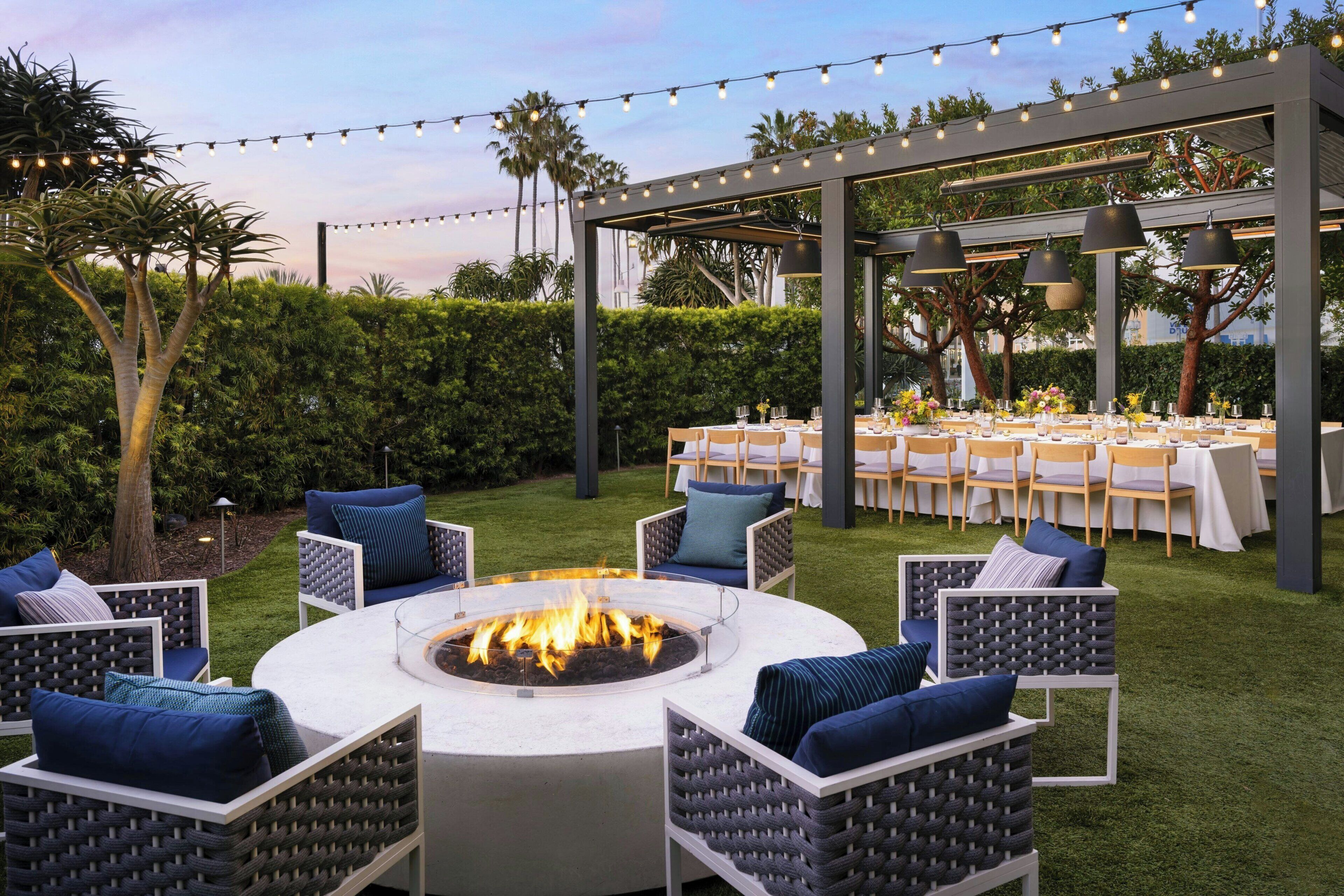An outdoor patio with a circular fire pit surrounded by wicker chairs, a covered dining area with tables set for a meal, and string lights overhead.