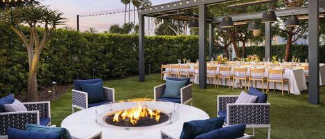 An outdoor patio with a circular fire pit surrounded by wicker chairs, a covered dining area with tables set for a meal, and string lights overhead.