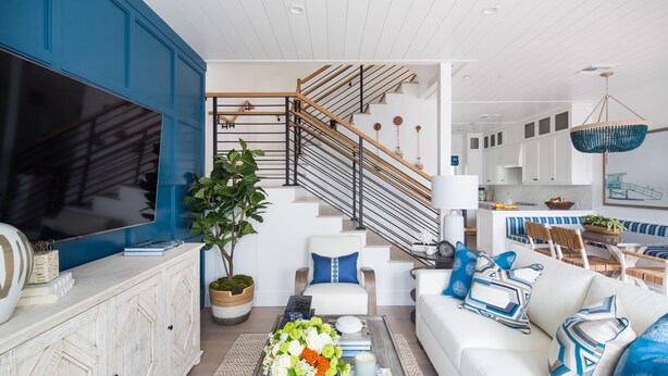 Ein modernes Wohnzimmer mit einem großen Flachbildfernseher, einem weißen Sofa, einem Couchtisch mit Büchern und Blumen sowie einer Treppe mit Holzbalkonen.