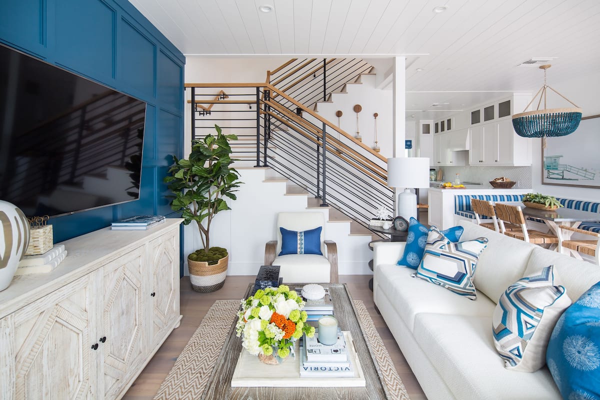 A modern living room with a large flat-screen TV, a white sofa, a coffee table with books and flowers, and a staircase with wooden railings.