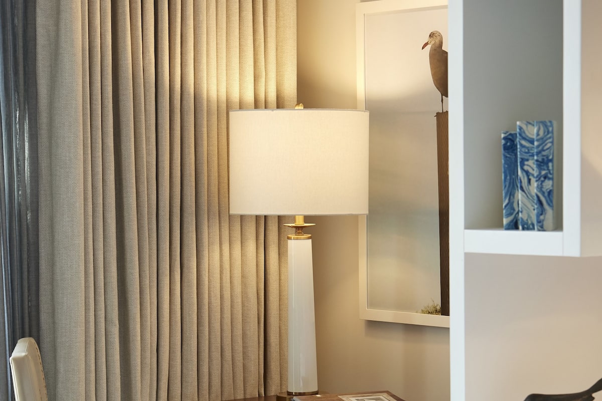 A hotel room with a bed, a desk with a lamp, a chair, and a bookshelf with decorative items.