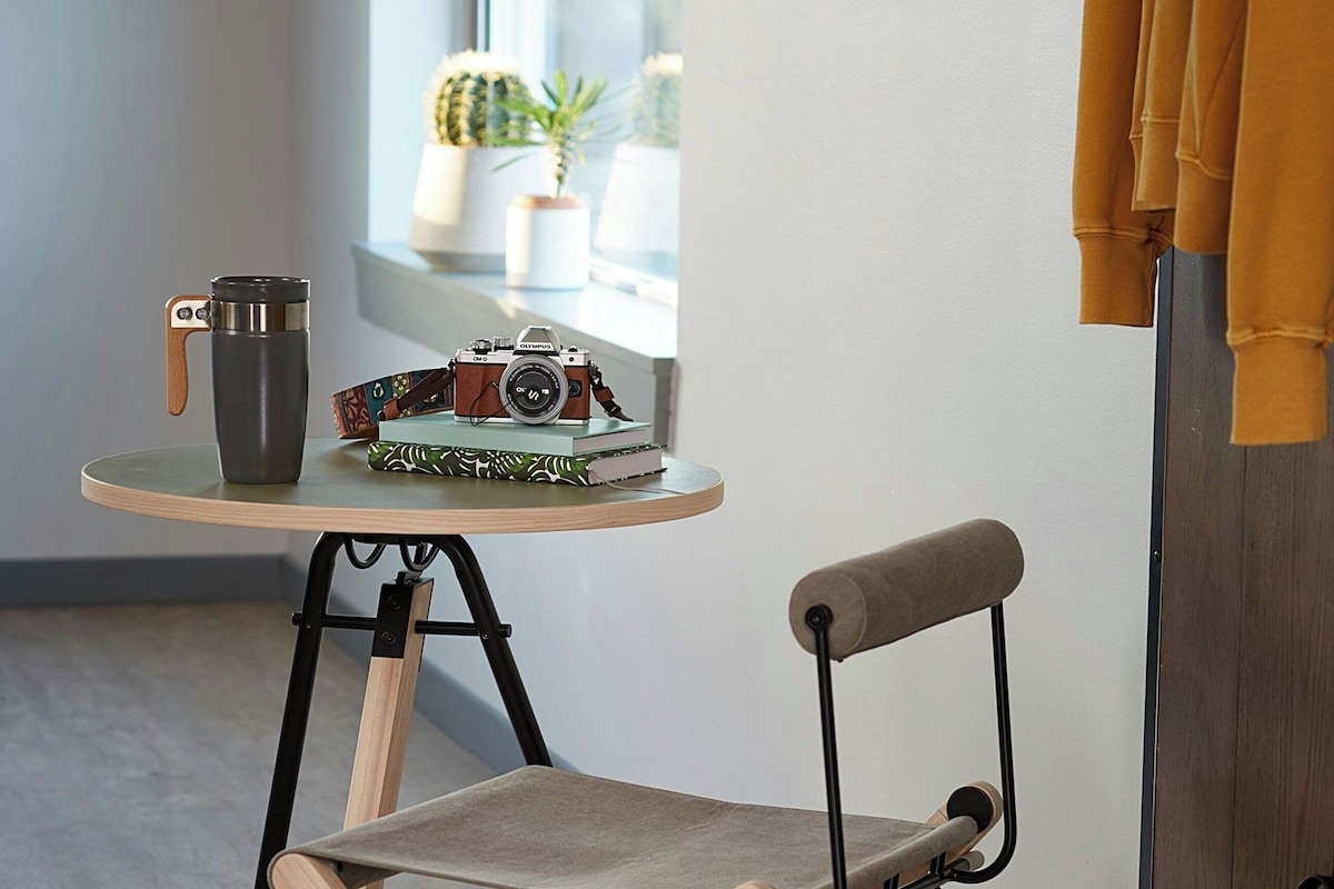 Une pièce de taille modeste, avec une chaise pliante, une petite table sur laquelle se trouvent un appareil photo et des livres, un sac à dos et un téléviseur fixé au mur.