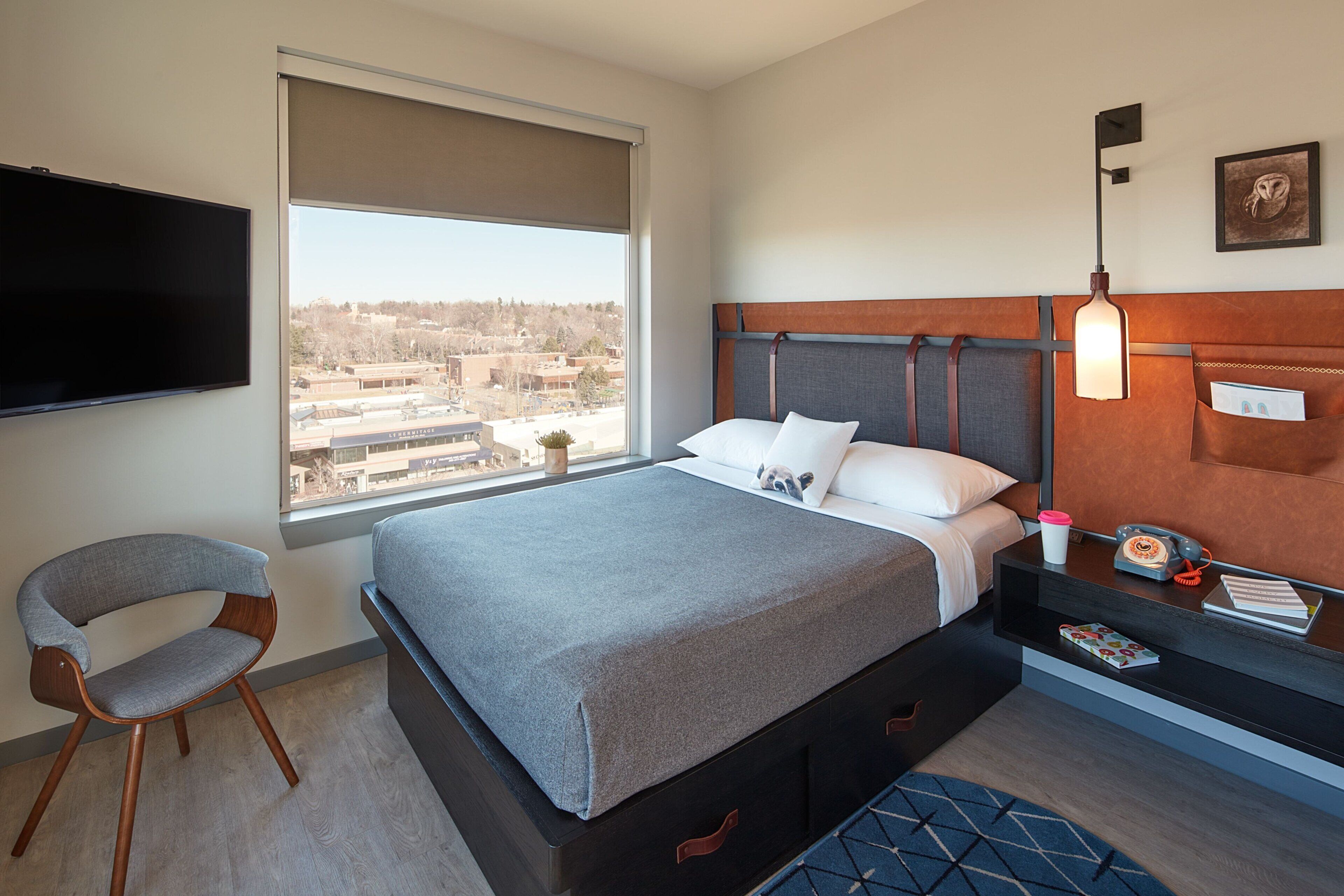 A hotel room with a bed, a chair, a bedside table with a lamp, a TV, and a window with a view of buildings.