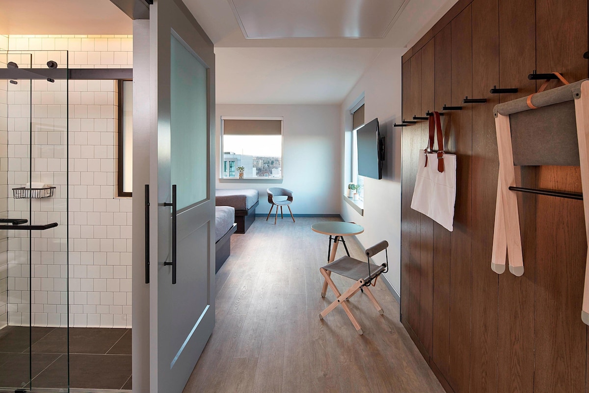Un couloir d’hôtel moderne comprenant une douche, un coin salon avec une table et des chaises, ainsi qu’une armoire en bois avec des vêtements suspendus.