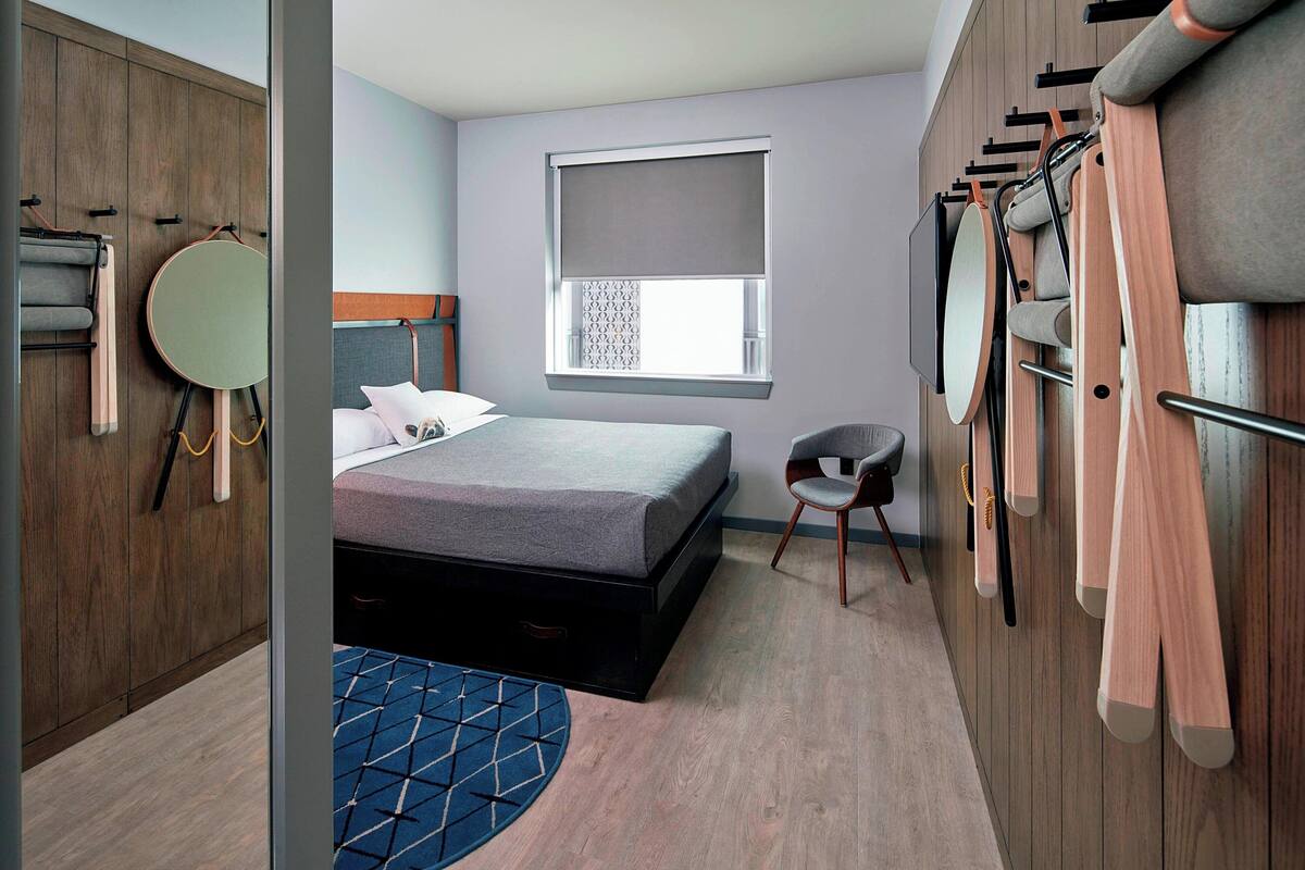 Een moderne slaapkamer met een bed, een raam met zonwering, een stoel en een kledingkast met daarin hangende kleding.