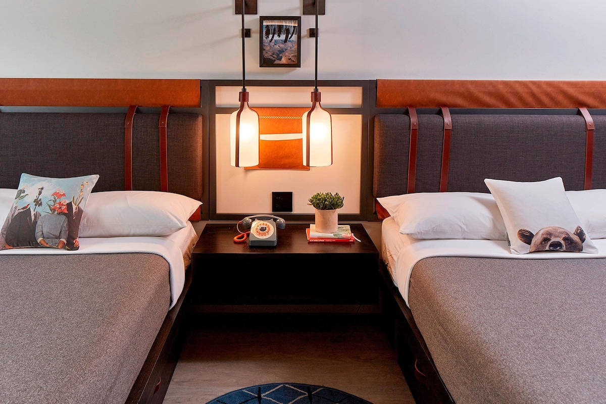 Une chambre d’hôtel avec deux lits, une table de chevet avec un téléphone et une plante, et un tableau au mur.