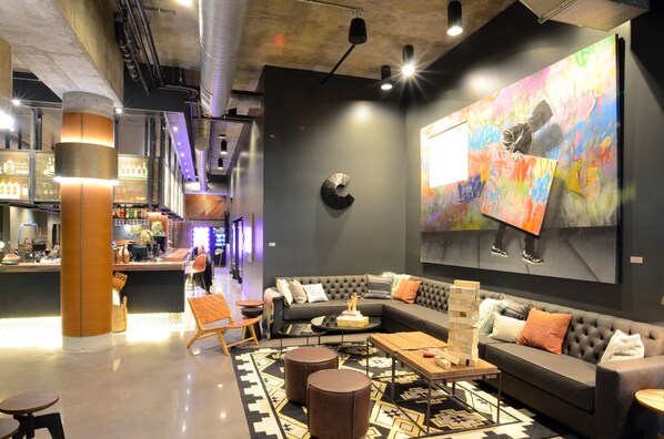 Un espacio moderno tipo lounge con un sofá, una mesa de centro y otomanas, una barra con un cantinero, y un cuadro abstracto grande en la pared.