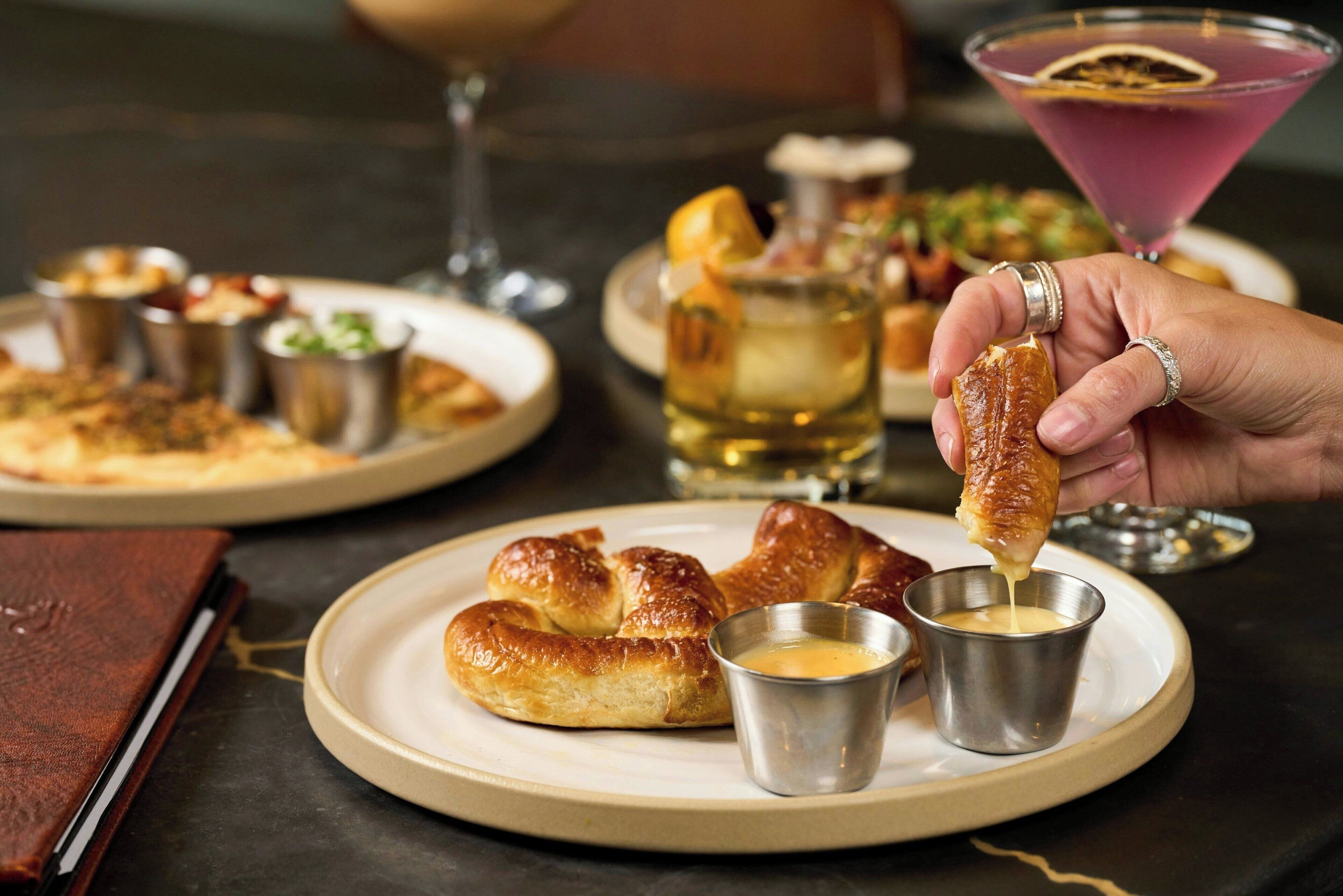 Un piatto con un croissant e due piccole tazze metalliche, probabilmente contenenti burro, su un tavolo con una bottiglia e un bicchiere da martini sullo sfondo.