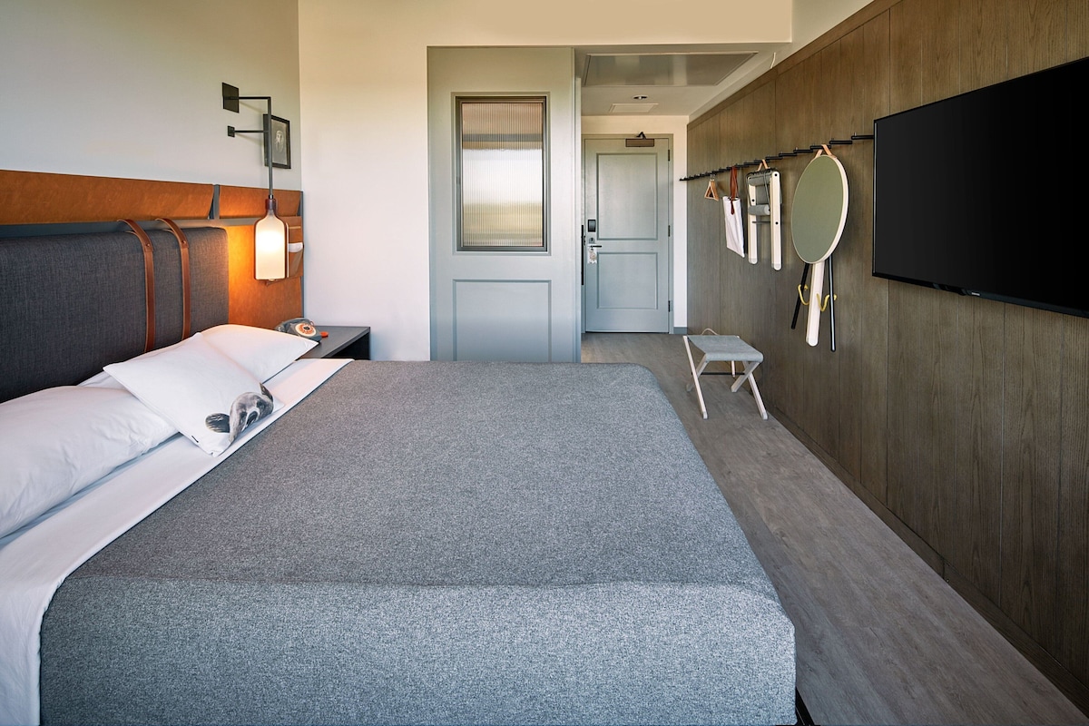 Une chambre d’hôtel avec un grand lit, une table de chevet avec une lampe, un téléviseur fixé au mur et une porte donnant sur une salle de bain.