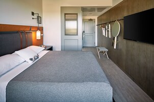 Una habitación de hotel con una cama grande, una mesita de noche con lámpara, un televisor montado en la pared y una puerta que da a un baño.