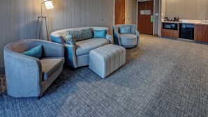 Habitación de hotel con alfombra gris, un sofá, una lámpara de pie, una kitchenette y una puerta.
