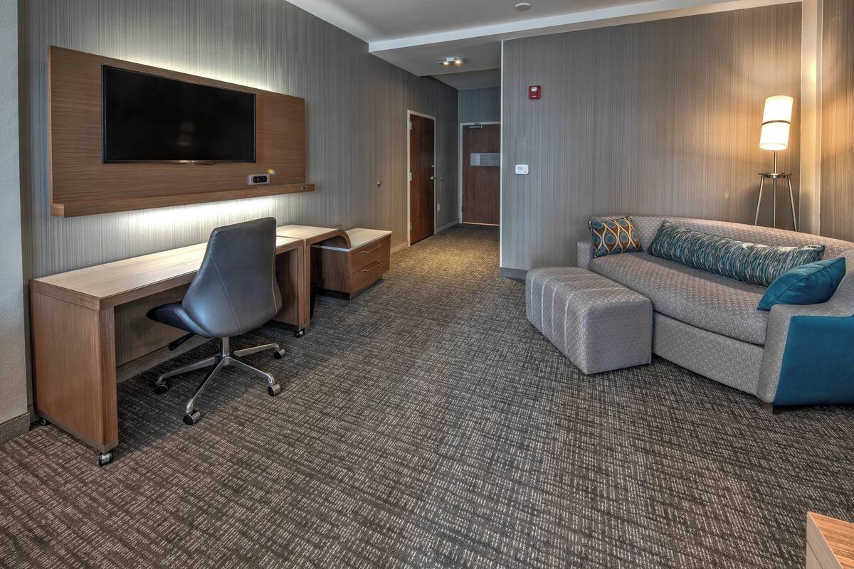 Ein Hotelzimmer mit Wandfernseher, Schreibtisch mit Stuhl, einer Couch mit Kissen und einer Stehlampe.