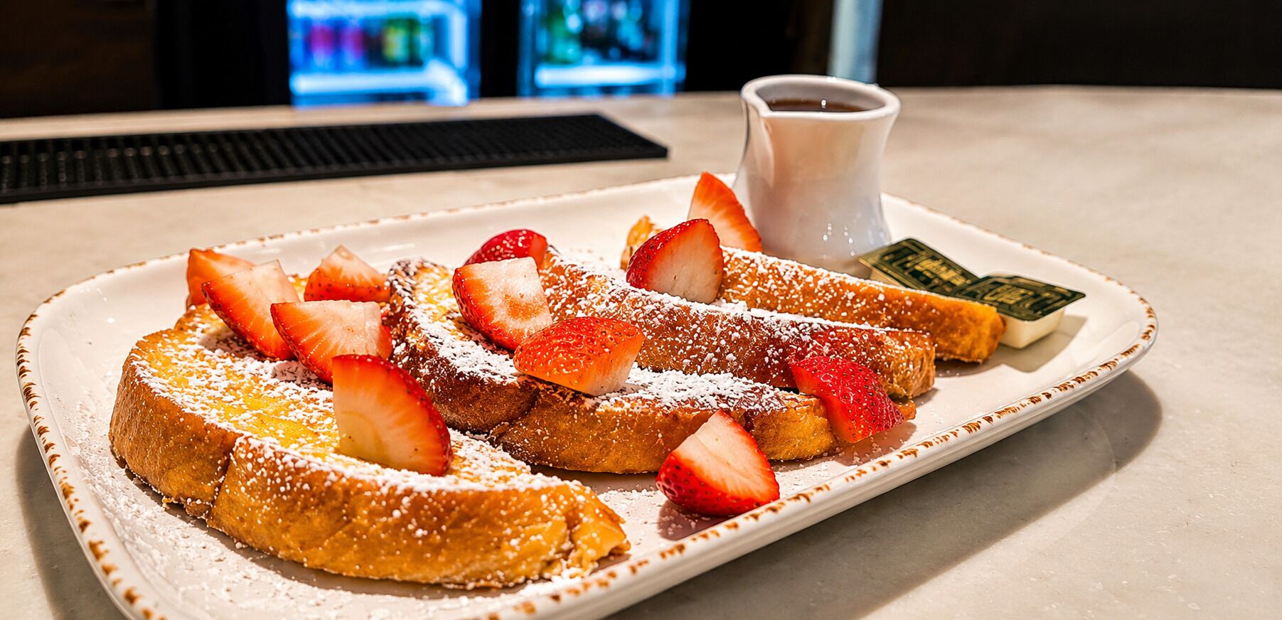 Une assiette de pain perdu garnie de fraises et saupoudrée de sucre glace, un petit pichet de sirop et un plat vert.