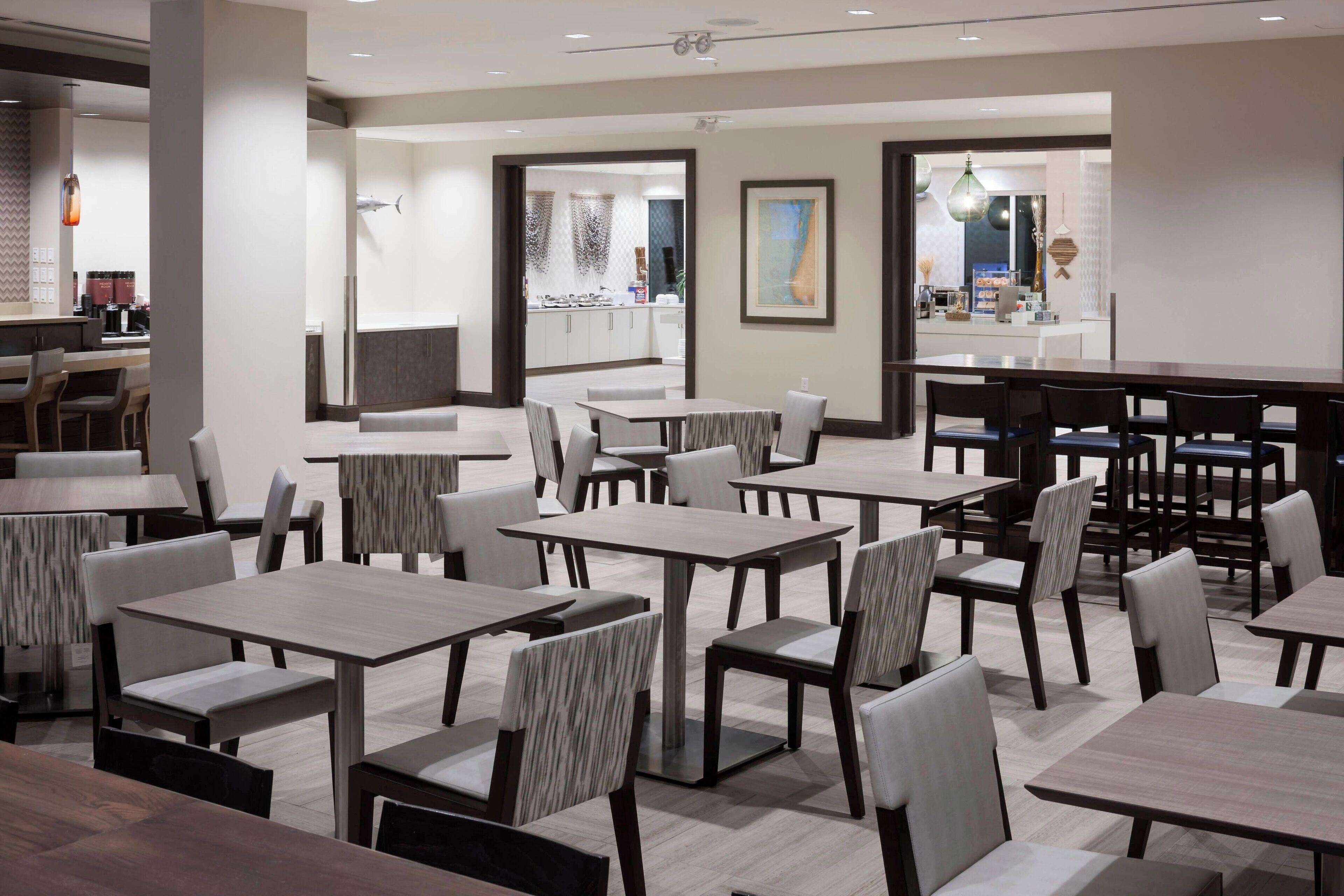 Un comedor de hotel moderno con varias mesas y sillas, una zona de bar y vistas a la cocina al fondo.