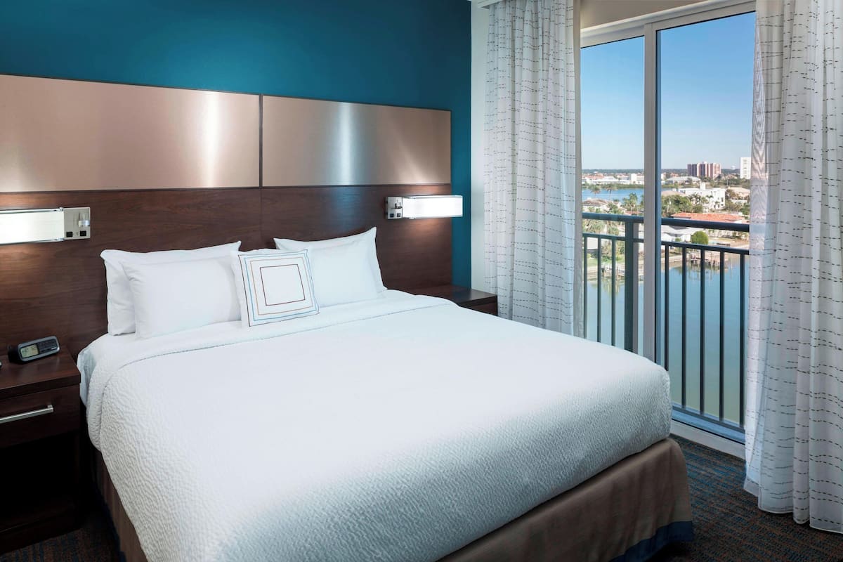 Une chambre d’hôtel avec un grand lit, une table de chevet, une lampe fixée au mur et une vue sur la ville depuis un balcon agrémenté de rideaux.