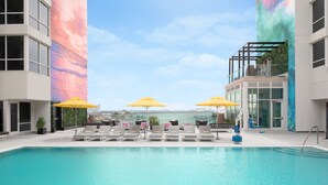 Zona de piscina en la azotea con sillones de relax y sombrillas amarillas, rodeada de edificios modernos con grandes ventanales.