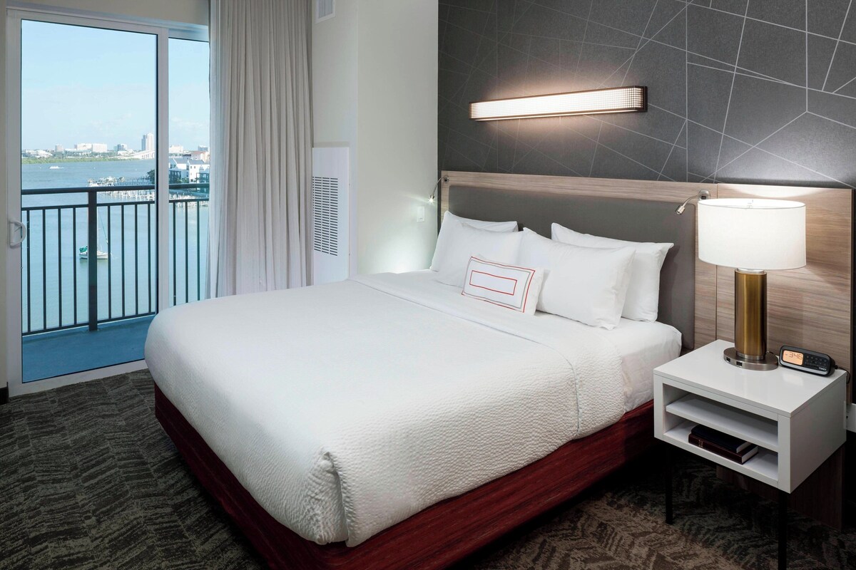 A hotel room with a large bed, a nightstand with a lamp, and a view of the city through the window.