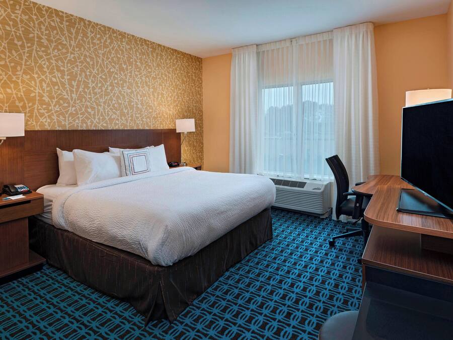 A hotel room with a large bed, two bedside tables with lamps, a desk with a flat-screen TV, and a patterned carpet.