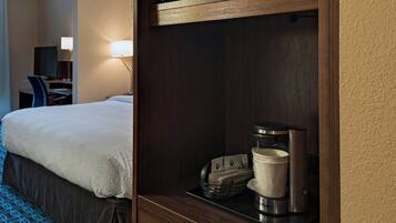 Quarto de hotel com cama, escrivaninha com luminária, micro-ondas e cafeteira em uma prateleira.