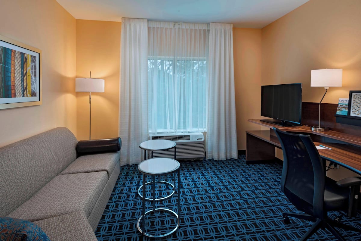 Une chambre d’hôtel avec un canapé, une petite table, un bureau avec une chaise, une télévision et une fenêtre avec des rideaux.