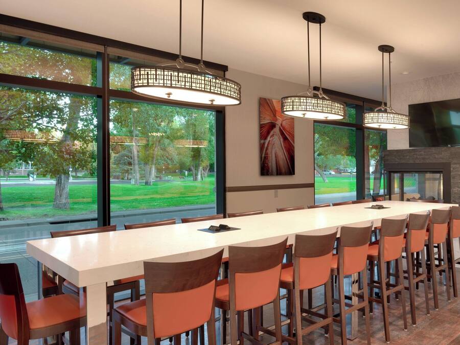 Un comedor moderno con una mesa larga, varias sillas, luces colgantes, ventanas amplias con vista a árboles y un televisor de pantalla plana montado en la pared.