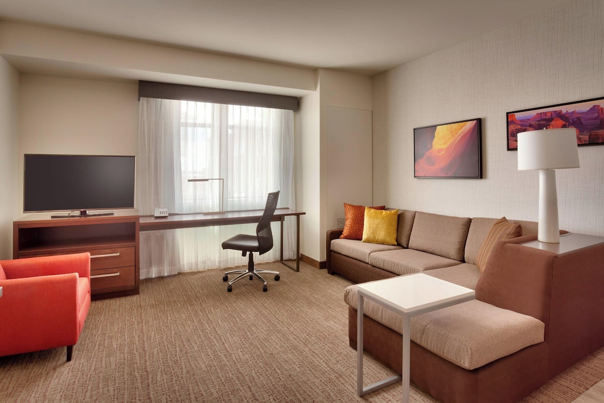 A hotel room with a large window, a flat-screen TV, a desk, a sofa, and a lamp.
