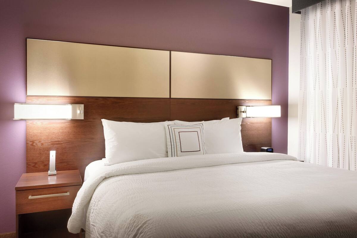 A neatly made bed with a wooden headboard, a bedside table with a lamp, and a nightstand with a phone.