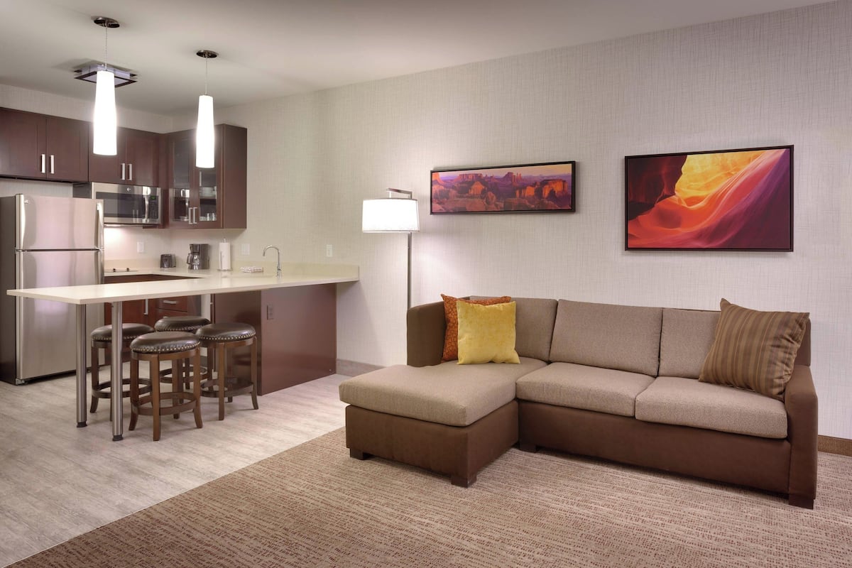 A modern kitchen with stainless steel appliances, a bar area with stools, a sofa with cushions, and two framed artworks on the wall.