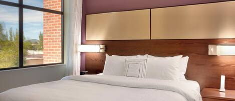 A hotel room with a large bed, a wooden headboard, a nightstand with a lamp, and a window with a view of greenery and a building.