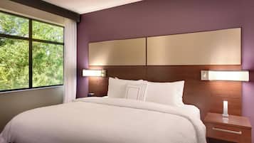 A neatly made bed with white linens, a wooden nightstand with a drawer, and a wall-mounted light fixture.
