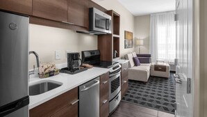 A modern kitchen with a built-in microwave, stainless steel appliances, and a coffee maker. There is a seating area with a sofa and a small table. - TownePlace Suites by Marriott Charlotte Fort Mill (Fort Mill)
