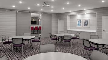 Un salón de conferencias con mesas y sillas redondas, un proyector y vista hacia una zona de cocina.