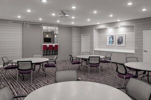 Un salón de conferencias con mesas y sillas redondas, un proyector y vista hacia una zona de cocina.