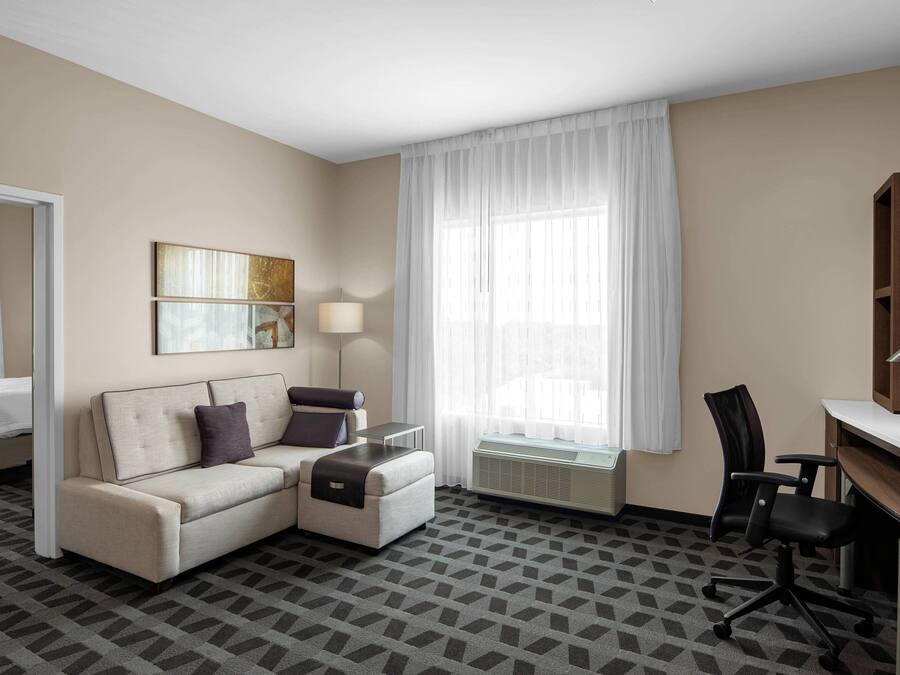 Habitación de hotel con sofá, escritorio, silla, lámpara y ventana con cortinas.