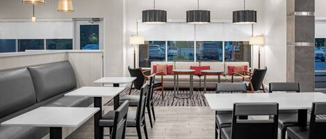 A modern dining area with a long bench, tables, and chairs, featuring pendant lighting and a view through large windows.