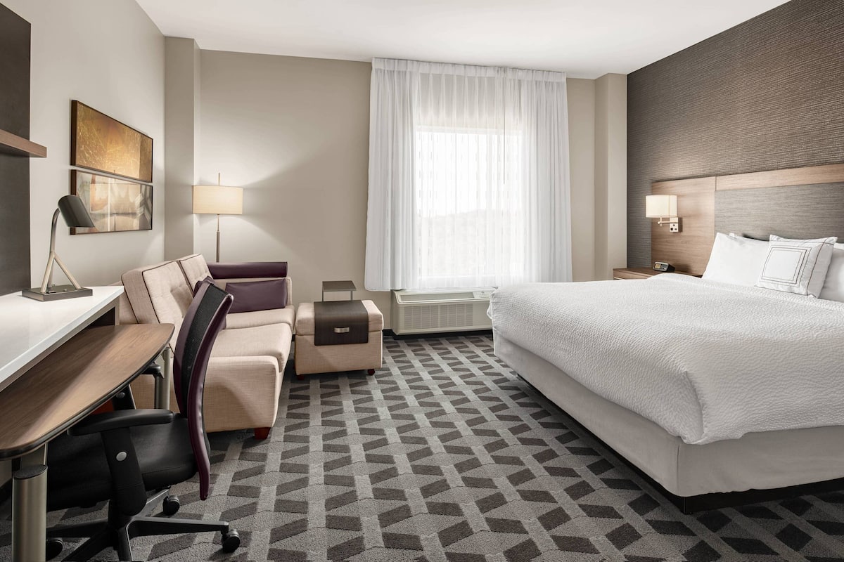 Una habitación de hotel con una cama grande, un escritorio con silla, un sofá, una lámpara y una ventana con cortinas traslúcidas.