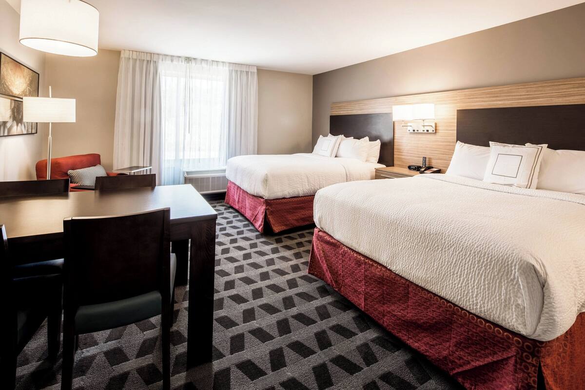 A hotel room with two beds, a desk, a chair, and a lamp.