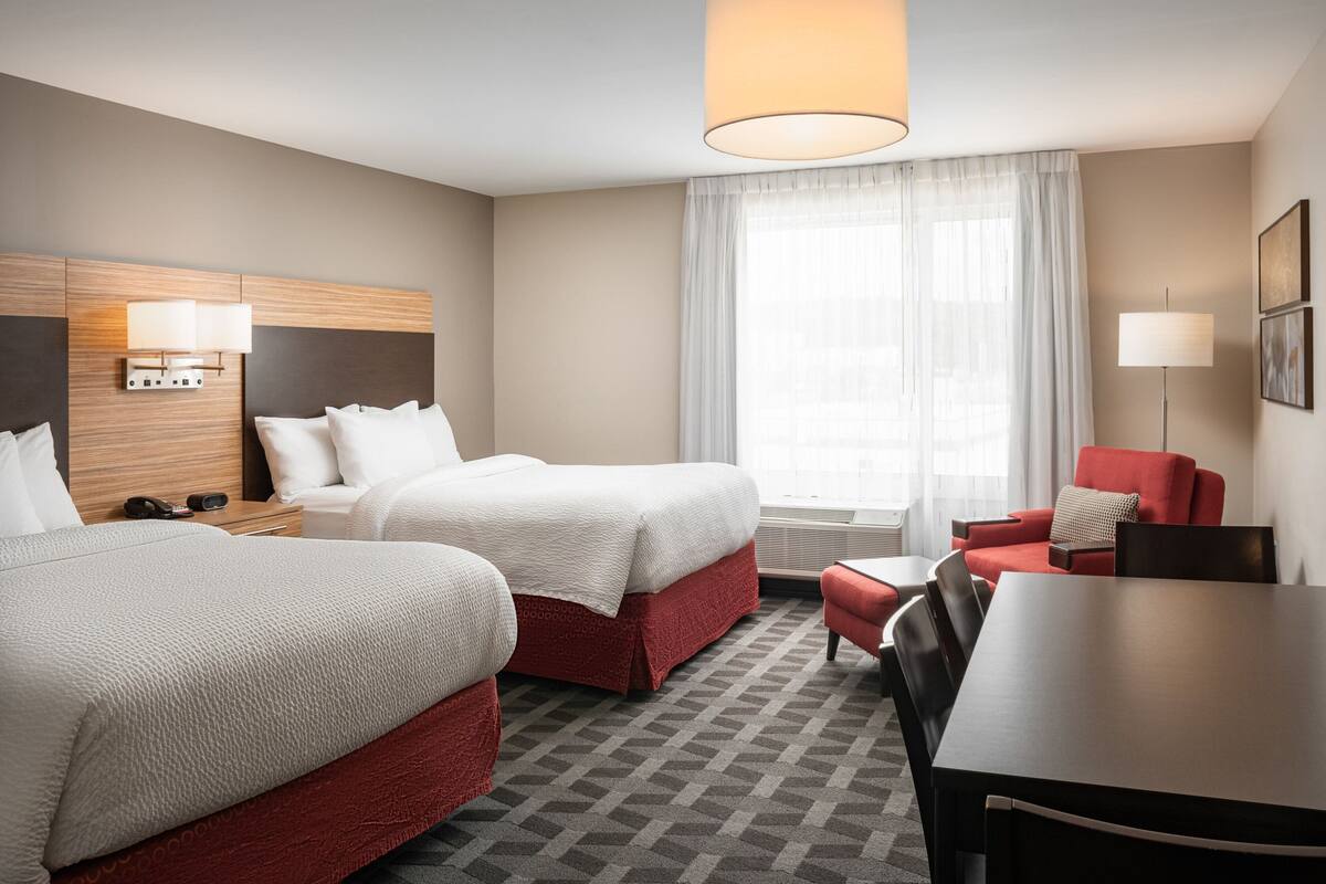 A hotel room with two beds, a dining table, a red armchair, and a large window with sheer curtains.