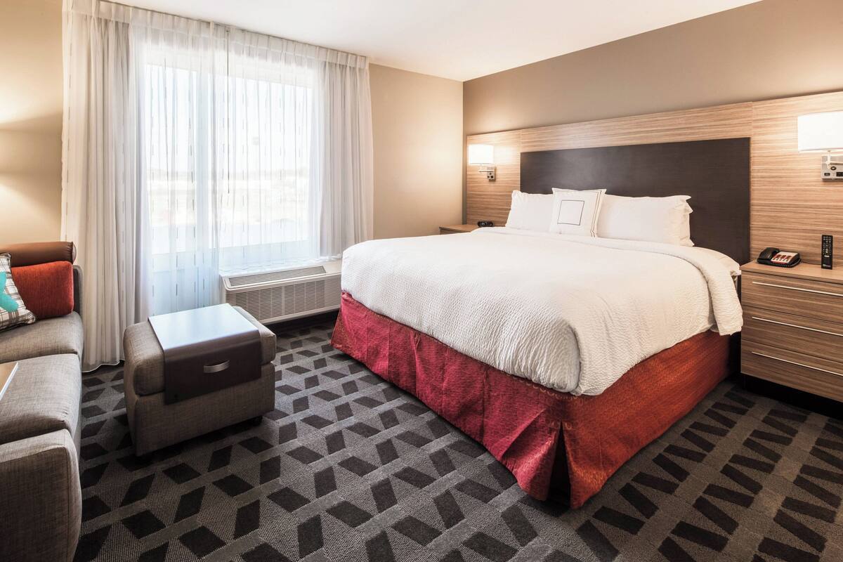 A hotel room with a large bed, a small sofa, a nightstand with a lamp, and a window with sheer curtains.