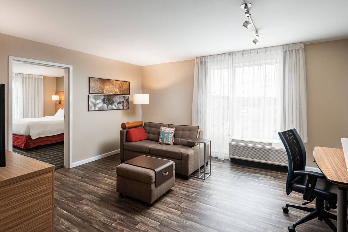 A modern hotel room with a wooden floor, a sofa, a small table, a desk, a chair, a bed, and a window with curtains.
