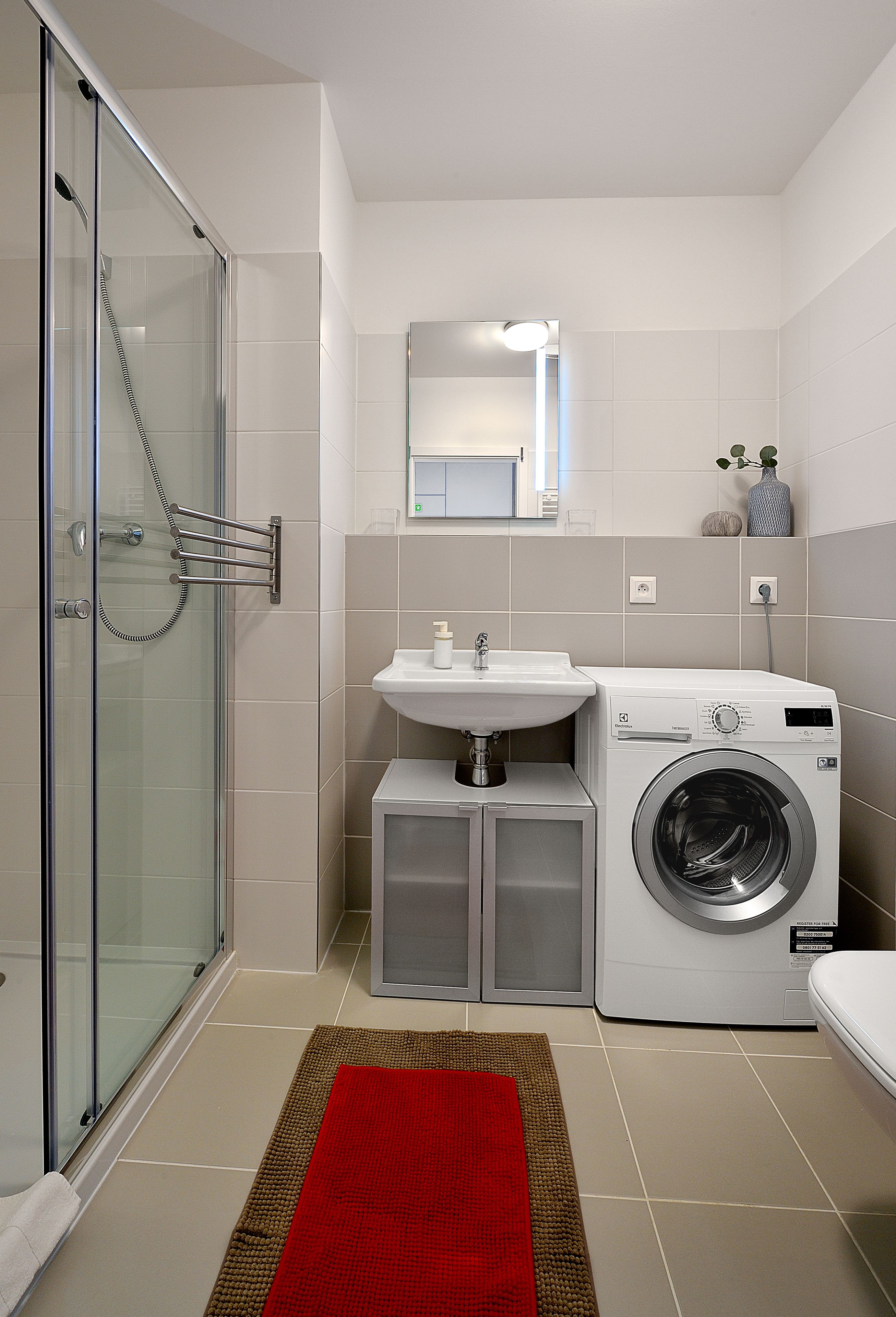 A compact bathroom with a glass shower enclosure, a sink, a washing machine, and a red rug.