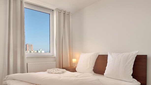 A neatly made bed with white linens, a wooden headboard, and a view of the city through a large window with white curtains. - Apollis Business & Budget by Ambiente (Bratislava)