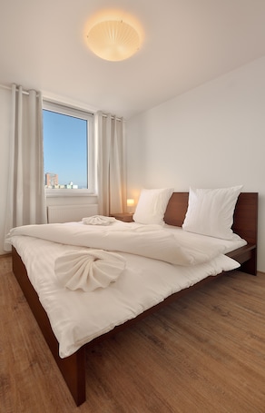 A neatly made bed with white linens, a wooden headboard, and a view of the city through a large window with white curtains.