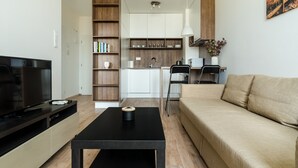 A modern living room with a flat-screen TV, a black coffee table, a beige sofa, and a kitchen area with white cabinets and wooden shelves. - Apollis Business & Budget by Ambiente (Bratislava)