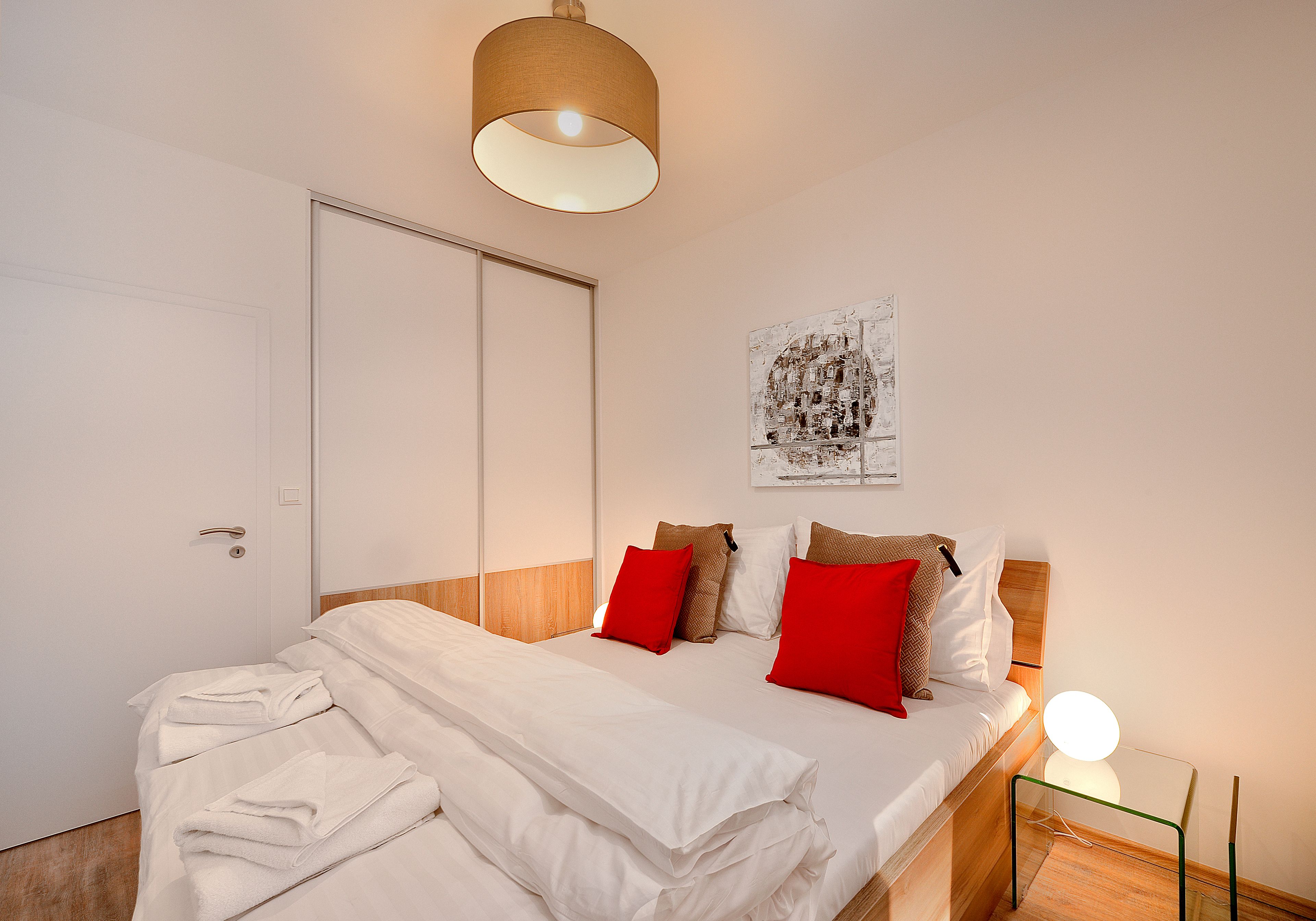 Ein ordentlich bezogenes Bett mit weißen Leinen und roten Kissen, ein hölzernes Kopfteil, ein an der Wand befestigtes Kunstwerk und ein Nachttisch mit einer Lampe.