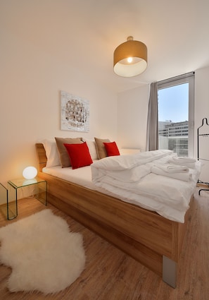 Een moderne slaapkamer met een houten bed, witte lakens, rode en bruine kussens, een glazen bijzettafeltje met een lamp, een zacht wit tapijt en een uitzicht op de stad door het raam.