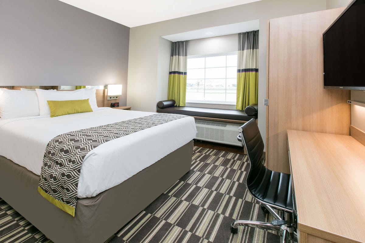 A hotel room with a large bed, a desk, a chair, a window with curtains, and a flat-screen TV mounted on the wall.